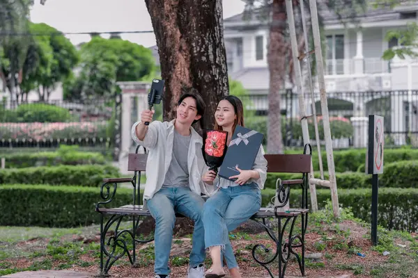 Asyalı çift, parktaki bankta eşine bir buket kırmızı çiçek ve hediye paketi sunan adamla samimi bir anın tadını çıkarıyor. Sıcak, açık hava manzarası aşkı, takdiri ve romantik kutlamayı yansıtıyor.