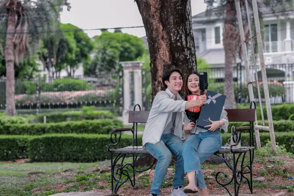 Asyalı çift, parktaki bankta eşine bir buket kırmızı çiçek ve hediye paketi sunan adamla samimi bir anın tadını çıkarıyor. Sıcak, açık hava manzarası aşkı, takdiri ve romantik kutlamayı yansıtıyor.