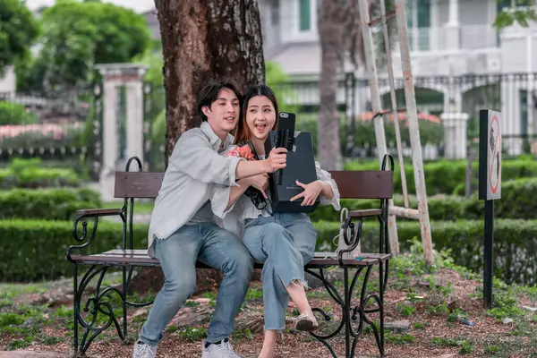 Asyalı çift, parktaki bankta eşine bir buket kırmızı çiçek ve hediye paketi sunan adamla samimi bir anın tadını çıkarıyor. Sıcak, açık hava manzarası aşkı, takdiri ve romantik kutlamayı yansıtıyor.