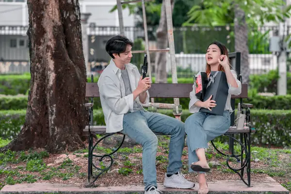 Asyalı çift, parktaki bankta eşine bir buket kırmızı çiçek ve hediye paketi sunan adamla samimi bir anın tadını çıkarıyor. Sıcak, açık hava manzarası aşkı, takdiri ve romantik kutlamayı yansıtıyor.