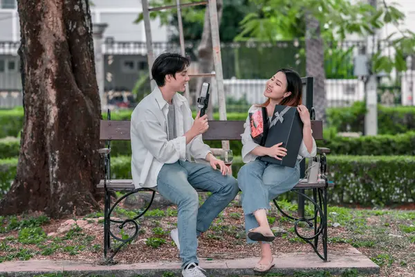 Asyalı çift, parktaki bankta eşine bir buket kırmızı çiçek ve hediye paketi sunan adamla samimi bir anın tadını çıkarıyor. Sıcak, açık hava manzarası aşkı, takdiri ve romantik kutlamayı yansıtıyor.