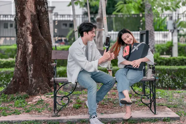 Asyalı çift, parktaki bankta eşine bir buket kırmızı çiçek ve hediye paketi sunan adamla samimi bir anın tadını çıkarıyor. Sıcak, açık hava manzarası aşkı, takdiri ve romantik kutlamayı yansıtıyor.