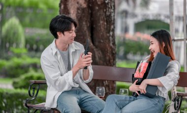 Asyalı çift, parktaki bankta eşine bir buket kırmızı çiçek ve hediye paketi sunan adamla samimi bir anın tadını çıkarıyor. Sıcak, açık hava manzarası aşkı, takdiri ve romantik kutlamayı yansıtıyor.