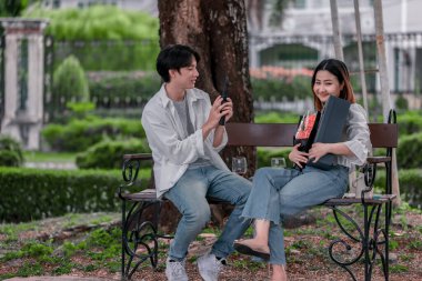 Asyalı çift, parktaki bankta eşine bir buket kırmızı çiçek ve hediye paketi sunan adamla samimi bir anın tadını çıkarıyor. Sıcak, açık hava manzarası aşkı, takdiri ve romantik kutlamayı yansıtıyor.