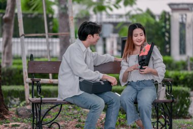 Asyalı çift, parktaki bankta eşine bir buket kırmızı çiçek ve hediye paketi sunan adamla samimi bir anın tadını çıkarıyor. Sıcak, açık hava manzarası aşkı, takdiri ve romantik kutlamayı yansıtıyor.