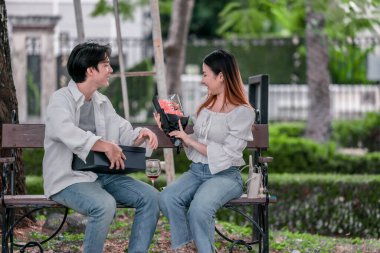 Asyalı çift, parktaki bankta eşine bir buket kırmızı çiçek ve hediye paketi sunan adamla samimi bir anın tadını çıkarıyor. Sıcak, açık hava manzarası aşkı, takdiri ve romantik kutlamayı yansıtıyor.