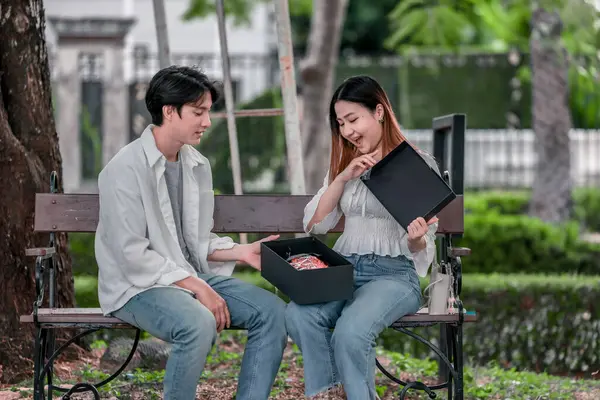 Asyalı çift, parktaki bankta eşine bir buket kırmızı çiçek ve hediye paketi sunan adamla samimi bir anın tadını çıkarıyor. Sıcak, açık hava manzarası aşkı, takdiri ve romantik kutlamayı yansıtıyor.