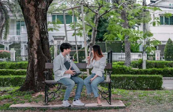 Asyalı çift, parktaki bankta eşine bir buket kırmızı çiçek ve hediye paketi sunan adamla samimi bir anın tadını çıkarıyor. Sıcak, açık hava manzarası aşkı, takdiri ve romantik kutlamayı yansıtıyor.