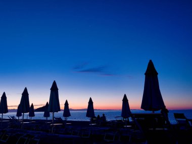 Şafak vakti kumlu bir sahilde şemsiyeler. Sıcak bir yaz gününde güneş şemsiyeler ve denizin arasından süzülürken.