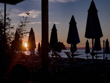 Şafak vakti kumlu bir sahilde şemsiyeler. Sıcak bir yaz gününde güneş şemsiyeler ve denizin arasından süzülürken.