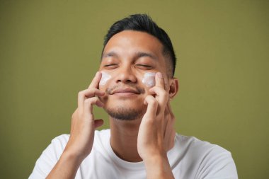 Potrait of asian man is applying moisturizing and anti aging cream on his face against isolated on green background