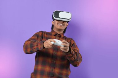 Potrait of young asian man smiling excited happy watching in vr headset pc gadget play pc game with joystick console isolated on purple background studio.