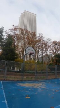 Geniş açı yavaş çekim sepeti ıslak ve kirli mavi basketbol sahasında sonbahar ağaçları ve arka planda beyaz bir kule ile Madrid 'de