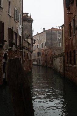 Her iki tarafında eski pencereleri olan, beyaz bulutlu gökyüzünü ve sudaki binaları yansıtan ve bulutlu bir sabahta ön planda tahta bir direği olan bir Venedik kanalı..