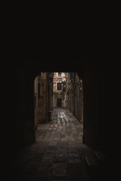 A shadowy stone alleyway with dim light in a narrow street of Venice, featuring a fire hydrant in the distance, on a cloudy day.