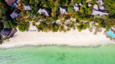 Aerial view of umbrellas, palms on the sandy beach of Indian Ocean at sunny day. Summer holiday in Zanzibar, Africa. Tropical landscape with palm trees, parasols, boats, yachts, blue water. Top view