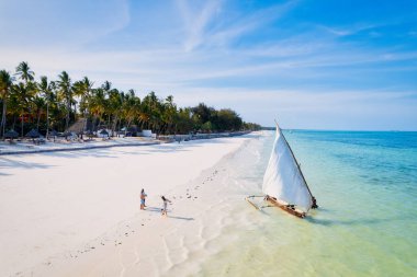 Relax on the white sand beach in Kiwengwa village on Zanzibar while admiring a Dhow catamaran sailboat gently gliding through the crystal-clear waters.