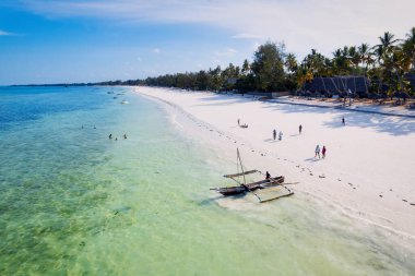Relax on the white sand beach in Kiwengwa village on Zanzibar while admiring a Dhow catamaran sailboat gently gliding through the crystal-clear waters.