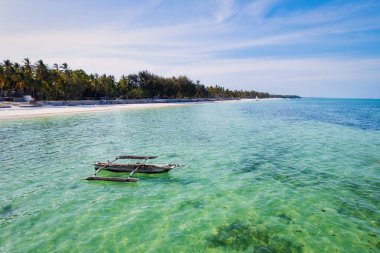 Relax on the white sand beach in Kiwengwa village on Zanzibar while admiring a Dhow catamaran sailboat gently gliding through the crystal-clear waters.