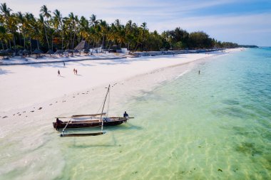 Relax on the white sand beach in Kiwengwa village on Zanzibar while admiring a Dhow catamaran sailboat gently gliding through the crystal-clear waters.