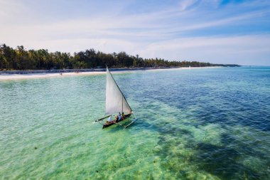 Relax on the white sand beach in Kiwengwa village on Zanzibar while admiring a Dhow catamaran sailboat gently gliding through the crystal-clear waters.
