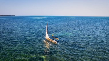Relax on the white sand beach in Kiwengwa village on Zanzibar while admiring a Dhow catamaran sailboat gently gliding through the crystal-clear waters.