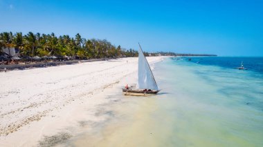 Zanzibar 'ın tropik cenneti güneş doğarken kumlu sahilde dinlenen balıkçı teknelerinin hava manzarasıyla canlanır. Manzarada berrak mavi sular, bereketli palmiye ağaçları ve uzakta bir yat bulunur..
