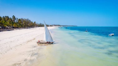 Zanzibar 'ın tropik cenneti güneş doğarken kumlu sahilde dinlenen balıkçı teknelerinin hava manzarasıyla canlanır. Manzarada berrak mavi sular, bereketli palmiye ağaçları ve uzakta bir yat bulunur..