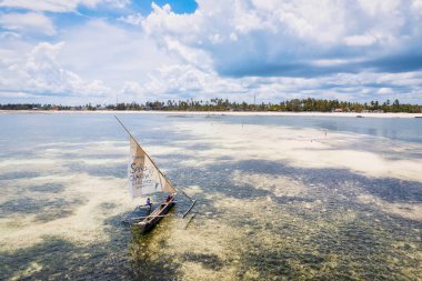 Zanzibar 'ın tropik cenneti güneş doğarken kumlu sahilde dinlenen balıkçı teknelerinin hava manzarasıyla canlanır. Manzarada berrak mavi sular, bereketli palmiye ağaçları ve uzakta bir yat bulunur..