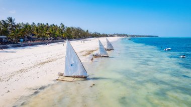 Zanzibar 'ın tropik cenneti güneş doğarken kumlu sahilde dinlenen balıkçı teknelerinin hava manzarasıyla canlanır. Manzarada berrak mavi sular, bereketli palmiye ağaçları ve uzakta bir yat bulunur..