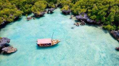 Zanzibar 'ın tropikal kıyı şeridinin havadan görünüşü güneş doğarken kumlu sahilde dinlenen balıkçı teknelerinin huzurlu görüntüsünü yakalar. Manzarada berrak mavi sular, bereketli palmiye ağaçları ve uzakta bir yat bulunur..