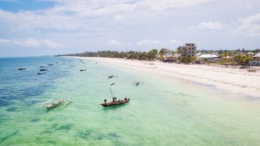 Zanzibar 'ın tropikal kıyı şeridinin havadan görünüşü güneş doğarken kumlu sahilde dinlenen balıkçı teknelerinin huzurlu görüntüsünü yakalar. Manzarada berrak mavi sular, bereketli palmiye ağaçları ve uzakta bir yat bulunur..