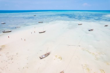 Zanzibar 'ın tropikal kıyı şeridinin havadan görünüşü güneş doğarken kumlu sahilde dinlenen balıkçı teknelerinin huzurlu görüntüsünü yakalar. Manzarada berrak mavi sular, bereketli palmiye ağaçları ve uzakta bir yat bulunur..