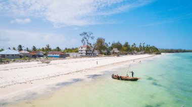 Zanzibar 'ın tropikal kıyı şeridinin havadan görünüşü güneş doğarken kumlu sahilde dinlenen balıkçı teknelerinin huzurlu görüntüsünü yakalar. Manzarada berrak mavi sular, bereketli palmiye ağaçları ve uzakta bir yat bulunur..