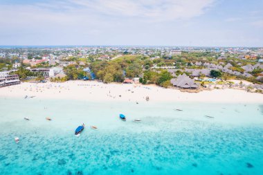 Zanzibar 'ın tropikal kıyı şeridinin havadan görünüşü güneş doğarken kumlu sahilde dinlenen balıkçı teknelerinin huzurlu görüntüsünü yakalar. Manzarada berrak mavi sular, bereketli palmiye ağaçları ve uzakta bir yat bulunur..