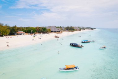 Zanzibar 'ın tropikal kıyı şeridinin havadan görünüşü güneş doğarken kumlu sahilde dinlenen balıkçı teknelerinin huzurlu görüntüsünü yakalar. Manzarada berrak mavi sular, bereketli palmiye ağaçları ve uzakta bir yat bulunur..