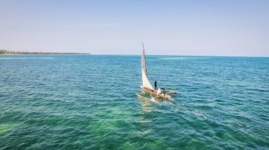 Zanzibar 'ın tropikal kıyı şeridinin havadan görünüşü güneş doğarken kumlu sahilde dinlenen balıkçı teknelerinin huzurlu görüntüsünü yakalar. Manzarada berrak mavi sular, bereketli palmiye ağaçları ve uzakta bir yat bulunur..