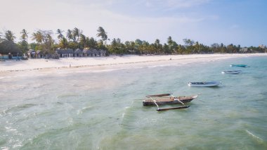 Zanzibar 'ın tropikal kıyı şeridinin havadan görünüşü güneş doğarken kumlu sahilde dinlenen balıkçı teknelerinin huzurlu görüntüsünü yakalar. Manzarada berrak mavi sular, bereketli palmiye ağaçları ve uzakta bir yat bulunur..