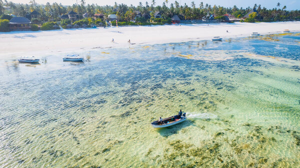 Experience the beauty of Zanzibar's tropical coast from a bird's eye view, with fishing boats resting on the sandy beach at sunrise. The top-down perspective showcases clear blue waters, green palm trees, and even a yacht.
