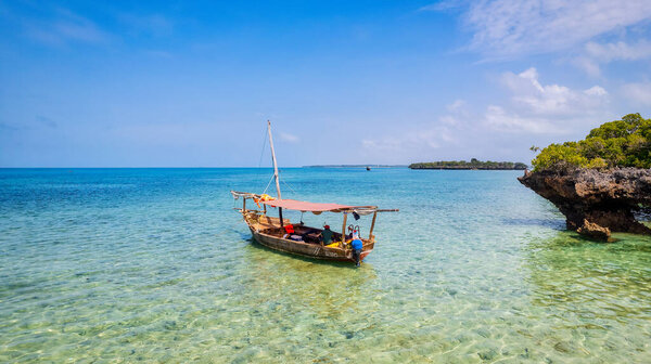 Take in the stunning scenery of Zanzibar's tropical coast from above, as fishing boats rest on the sandy beach at sunrise. The view from the top reveals clear blue waters, green palm trees, and even a yacht in the distance.