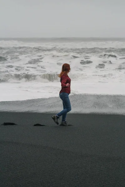 黒いアイスランドのビーチの風光明媚な写真に沿って歩く女性 背景に嵐の海を持つ人の写真 高品質の壁紙 旅行ブログ 記事のための写真のコンセプト — ストック写真