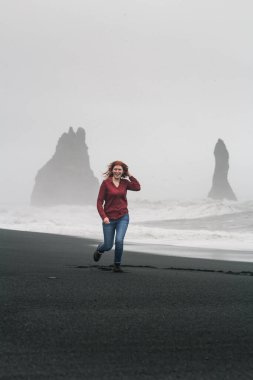 Smiling woman running along black beach scenic photography. Picture of person with rock columns on background. High quality wallpaper. Photo concept for ads, travel blog, magazine, article
