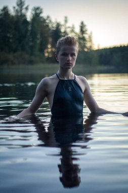 Close up attractive woman in lake at sunset portrait picture. Closeup front view photography with fir forest on background. High quality photo for ads, travel blog, magazine, article