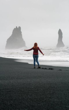 Lady enjoying view of rocks on black Iceland beach scenic photography. Picture of person with misty sea on background. High quality wallpaper. Photo concept for ads, travel blog, magazine, article