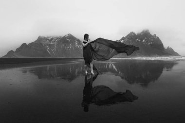 Elegant lady with long waving cape on beach monochrome scenic photography. Picture of person with hills on background. High quality wallpaper. Photo concept for ads, travel blog, magazine, article