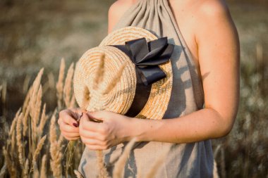 Close up pretty female hands holding straw hat with ribbon concept photo. Front view photography with grainfield on background. High quality picture for wallpaper, travel blog, magazine, article