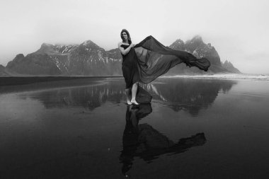 Lady with long chiffon cape on sea beach monochrome scenic photography. Picture of person with hills on background. High quality wallpaper. Photo concept for ads, travel blog, magazine, article