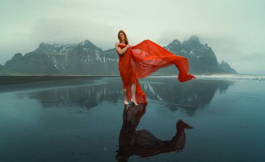 Redhead model in red dress on Reynisfjara beach scenic photography. Picture of person with hills on background. High quality wallpaper. Photo concept for ads, travel blog, magazine, article