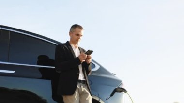 Akıllı telefon kullanan işadamı ve beklemede olan güç kaynağı arabadaki bataryayı şarj etmek için elektrikli araçlara bağlanıyor. Bir elektrikli arabayı şarj istasyonundan şarj etmek.