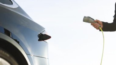 Unrecognizable man plugging electric car from charging station. Male plugging in power cord to electric car using app on smartphone. Businessman charging electric car at outdoor charging station